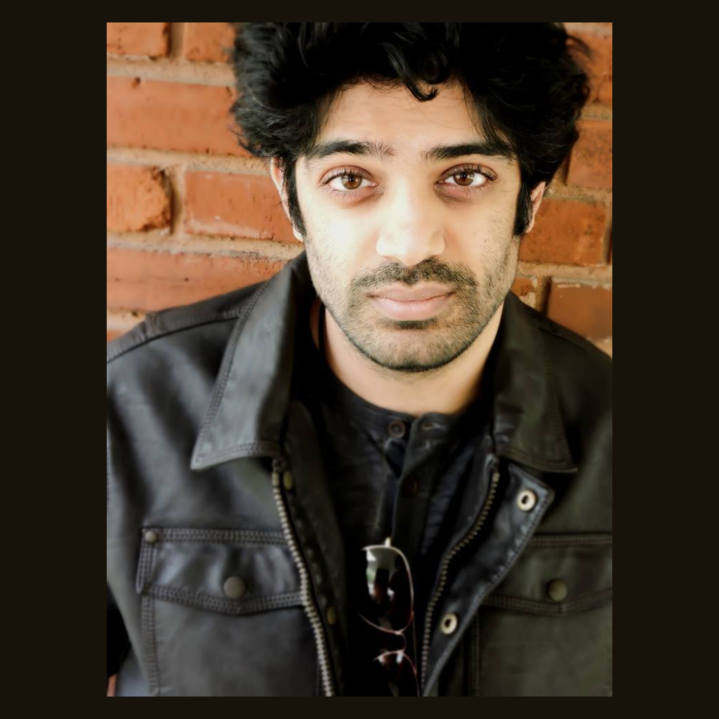 A portrait of author R. R. Virdi. He is an Indian-American man with short black hair and beard stubble, standing in front of a brick wall, wearing a black leather jacket, black shirt, and his sunglasses are hanging off the collar of the shirt. The look is smolder, even if he denies it. 